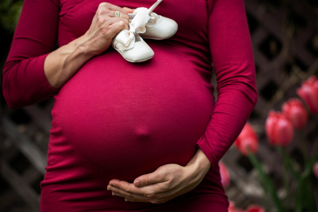 pexels photo 2617210 2617210 Pregnant woman holding baby shoes on belly in garden setting.